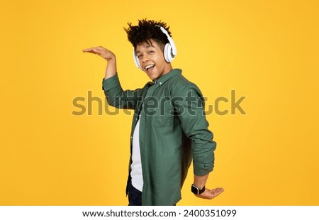 Cool handsome stylish young black guy hipster listening to music and dancing, smiling at camera, using brand new wireless stereo headphones, isolated on yellow studio background. Entertainment