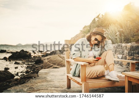 Cool happy beautiful girl woman sitting in a deck chair by the sea, stylish trendy outfit with hat and sunglasses, summer lifestyle sunny portrait.