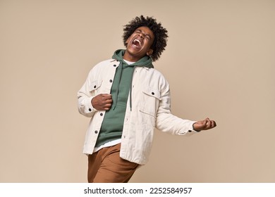 Cool happy hipster African American teen guy pretending playing guitar having fun isolated on beige background. Crazy funky ethnic generation z teenager fashion model screaming and dancing.