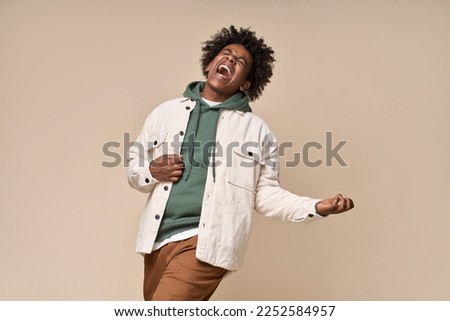 Cool happy hipster African American teen guy pretending playing guitar having fun isolated on beige background. Crazy funky ethnic generation z teenager fashion model screaming and dancing.