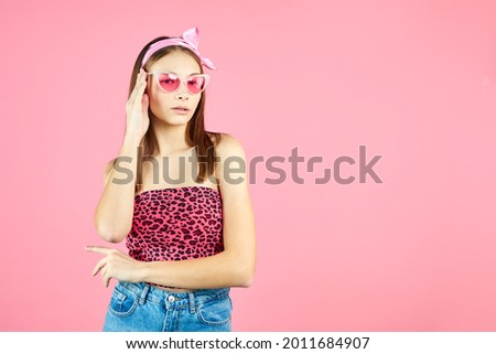 Cool hipster girl wearing stylish glasses. Positive young woman looking at camera on pink background. Caucasian female university student posing.