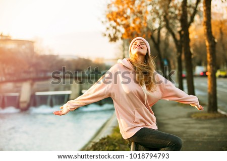 cool hipster young girl in pink hoodie standing by a river in the city enjoying the sun of autum or springtime