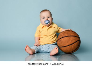 Cool Infant Baby Boy With Pacifier And In Jeans And Yellow Shirt Is Sitting On The Floor Holding Basket Ball On Background With Free Copy Space