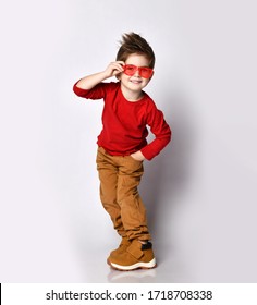 Cool Kid Boy With Stylish Hairstyle In Red Jumper, Brown Pants Stands Side To Us With His Hand In Pocket And Holding His Modern Sunglasses. Childhood, Fashion, Advertising