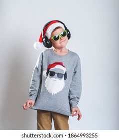 Cool Little Santa Claus Listening To Music With Headphones. Child Boy In Sunglasses And Stylish Christmas Sweater Isolated On Grey. Portrait Of School Kid On Holidays.