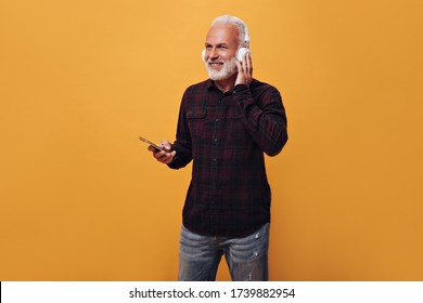 Cool Man In Plaid Shirt And Jeans Poses In Headphones On Beige Background. Attractive Adult In White Headphones With Phone In His Hands Having Fun