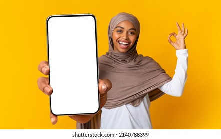 Cool mobile app. Happy muslim black lady showing smartphone with blank screen and okey gesture over yellow background. Woman holding mobile phone with empty screen, mockup