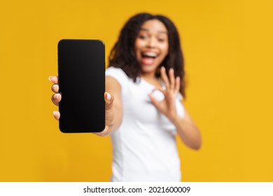Cool mobile app. Joyful black woman showing smartphone with blank screen and okey gesture over yellow background, selective focus. African american lady holding mobile phone with empty screen. Mockup