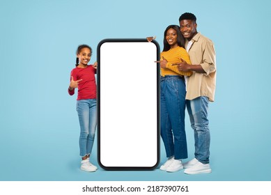 Cool Mobile Application. Happy Black Family Pointing At Big Giant Smartphone With Mockup, Promoting App Or Website Over Blue Background. Full Body Length