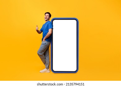 Cool Mobile Offer. Excited casual guy leaning on big smartphone with blank empty white display, showing thumb up gesture, recommending great new app or website, mock up, full body length, yellow wall