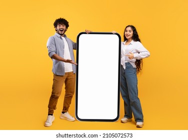 Cool mobile offer. Happy indian couple pointing at big cellphone with white screen for mockup, demonstrating free space for phone application or website design, yellow background