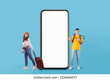 Cool Mobile Offer For Travellers. Couple of tourists presenting big giant cell, excited guy with backpack pointing at huge white blank screen, lady holding bag on blue studio wall, full body length