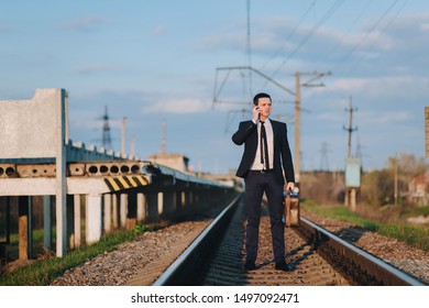 A cool and modern male businessman in a suit with a suitcase is walking along the paths from the train and talking on the phone. Business walk and serious conversation. Late for the train.
