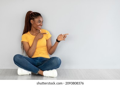 Cool offer. Happy young black woman sitting cross legged on floor, pointing aside at copy space against grey studio wall. Positive African American woman offering place for ad or promotional text