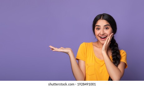 Cool Offer. Portarit of excited young indian woman holding hand, showing free copy space on palm and touching cheek standing isolated over purple studio background. Lady indicating place for advert