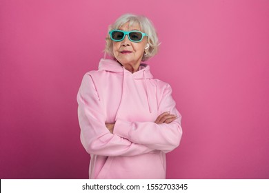 Cool Old Woman In Funny Green Sunglasses Posing On Camera. Hands Crossed. Wear Pink Hoody. Stylish Modern Grandma. Isolated Over Pink Background