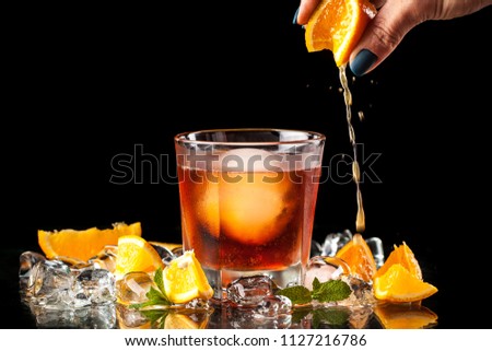 A cool orange alcohol cocktail with an ice ball in a glass on the bar. on the table, ice, orange, mint. The barman squeezes the juice into the glass with his hand. Concept image for bars, cafes.