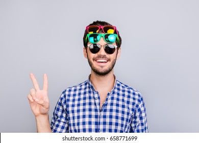 Cool! Playful young brunette bearded man in three pairs of bright colorful glasses and checkered casual shirt is fooling around, posing and shows v sign, smiling on light background