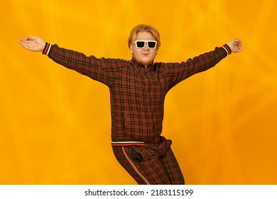 A Cool Plump Guy With Red-hair In Stylish Sunglasses And A Casual Sports Suit Dances A Street Dance On A Bright Yellow Background. Youth Culture. Hip-hop Style.