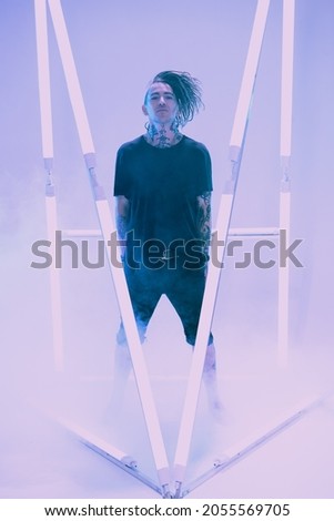 Cool punk rock star with dreadlocks in black clothes poses among neon lamps with his hands in his pockets surrounded by haze. Space punk rock music. Youth alternative culture. Full length portrait.