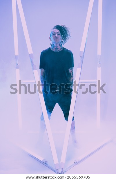 Cool punk rock star with dreadlocks in black\
clothes poses among neon lamps with his hands in his pockets\
surrounded by haze. Space punk rock music. Youth alternative\
culture. Full length\
portrait.