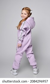 Cool Redhead Kid Girl In Pink Jumpsuit With Hood And Reflective Safety Stripes, Stands Back To Camera And Turned With Legs Wide Apart Over Light Background