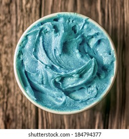 Cool Refreshing Turquoise Blue Ice Cream In A Takeaway Tub Standing On An Old Textured Wooden Table, Overhead View In Square Format