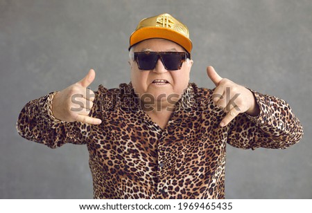 Cool retired gangsta granddad flexing to rap music. Portrait of funny weird active rich senior man in baseball cap, trendy eyeglasses and leopard patterned party shirt posing and having fun in studio