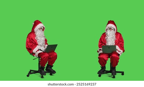 Cool saint nick using laptop on chair, sitting against full body greenscreen backdrop in studio. Father christmas working with modern portable pc, browsing online website in red costume.
