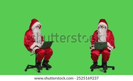 Cool saint nick using laptop on chair, sitting against full body greenscreen backdrop in studio. Father christmas working with modern portable pc, browsing online website in red costume.