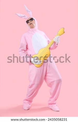 Cool senior man in Easter bunny costume playing paper guitar on pink background