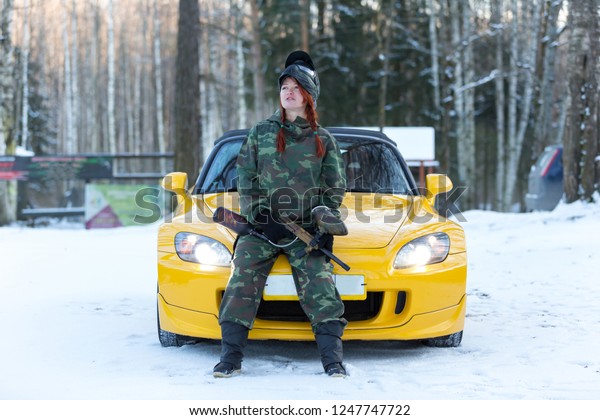 Cool sexy red hair girl in camouflage\
with paintball gun sitting on sport car\
bonnet