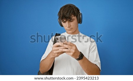 Cool and smart young hispanic teenager, a relaxed student, rocking out with headphones while typing on his smartphone, standing casual against an isolated blue background.