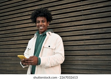 Cool smiling young African American guy holding mobile phone standing at wooden city wall. Happy stylish hipster teenager using cell outdoors, looking at camera with cellphone, typing on smartphone. - Powered by Shutterstock