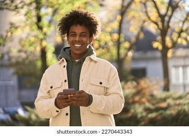 Cool smiling young African American guy holding mobile phone standing with smartphone at big city sunny street. Happy stylish hipster gen z teen using cellphone device outdoors, looking at camera. - Powered by Shutterstock