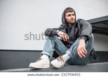 Cool street style man hipster in fashion casual wear with trendy rock leather jacket, hoodie, jeans and white sneakers sitting near a white wall on the street