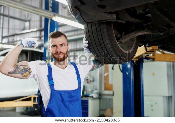 Cool strong repairman young professional\
technician mechanic man 20s wears denim blue overalls white t-shirt\
stand near car lift show hand biceps muscles work in vehicle repair\
shop workshop indoors
