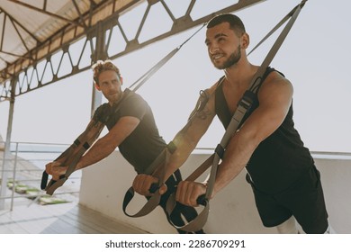 Cool strong young sporty athletic toned fit sportsman man in sport clothes warm up suspended training work out with TRX trainer at sunrise sun dawn over sea beach seaside outdoor in summer day morning