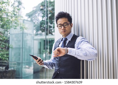 Cool Stylish Asian Businessman In Glasses And Vest Suit Checking Time On Wristwatch Standing In Hall Of Office Building With Smartphone In Hand