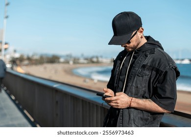 Cool stylish guy with black cap with sunglasses in denim jacket holding smartphone and sitting on promenade in Barcelona, ​​Spain - Powered by Shutterstock