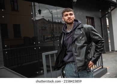 Cool Stylish Young Hipster Guy With Hair, A Beard, And A Tattoo On His Neck Wearing Fashion Casual Clothes With A Leather Rock Jacket, Hoodie, And Jeans Walks Near A Black Modern Building