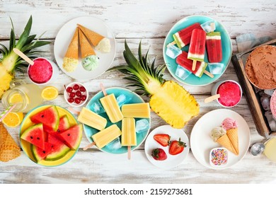 Cool summer foods table scene. Collection of ice cream, popsicles and fruit. Overhead view on a rustic white wood background.