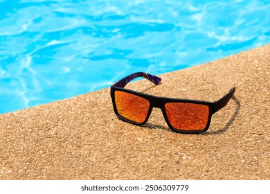 Cool sunglasses on the gravel poolside floor at swimming pool for summer vacation and holiday summer concept, Black sunglasses with orange lenses on tropical season in sunny day.