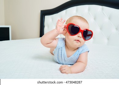 Cool Super Child.  Funny Baby Boy In Solar Glasses Of Red Heart Lies On A White Bed In A Room Inside The Building, Holds The Hand Points. 