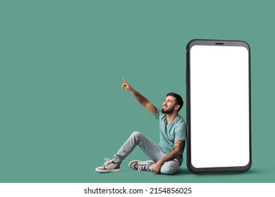 Cool tattooed young man sitting near big smartphone and pointing at something on green background
