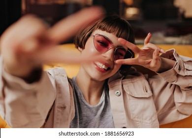 Cool teen girl in pink glasses showing peace signs inside. Happy short haired woman in light clothes smiling and sitting on yellow couch in cafe..