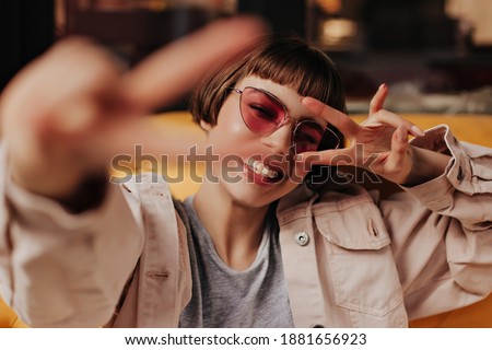 Cool teen girl in pink glasses showing peace signs inside. Happy short haired woman in light clothes smiling and sitting on yellow couch in cafe..