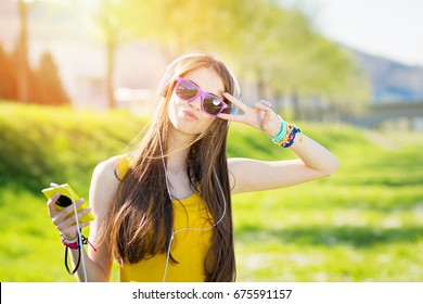 Cool Teenage Girl With Smart Phone And Headphones Outdoors In Summer Posing. Young Woman In Sunglasses Making Funny Faces And Posing. Natural Lighting, Retouched, Vibrant Colors.