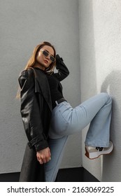 Cool Trendy Hipster Street Model Beauty Girl With Vintage Sunglasses In A Trendy Black Leather Jacket With A T-shirt And Fashion High Waisted Flared Jeans With Sneakers Poses Near A Wall In The City