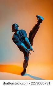 Cool trendy stylish professional dancer man with sunglasses and a cap in fashion black clothes with sneakers dances and lifts leg up in a creative colored studio with warm orange and cold neon light - Shutterstock ID 2278035731
